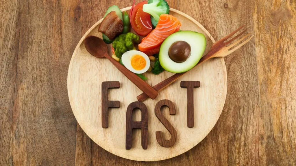 A wooden plate with keto-friendly foods like avocado, salmon, eggs, and vegetables, arranged like a clock, with wooden utensils pointing as clock hands. The word 'FAST' is displayed at the bottom, symbolizing intermittent fasting and its connection to the keto lifestyle